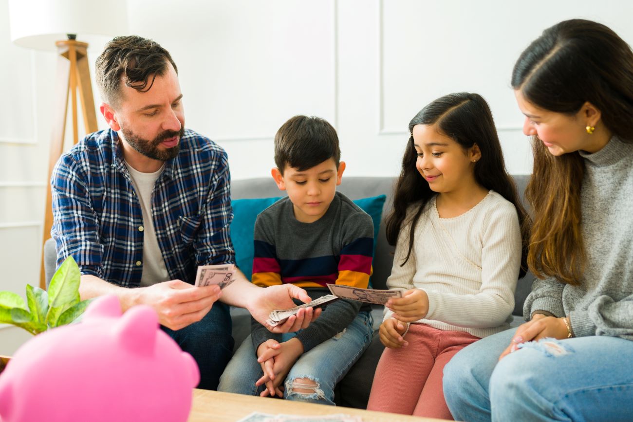 Family of man, boy, girl, woman sitting on couch and man hands allowance money to kids