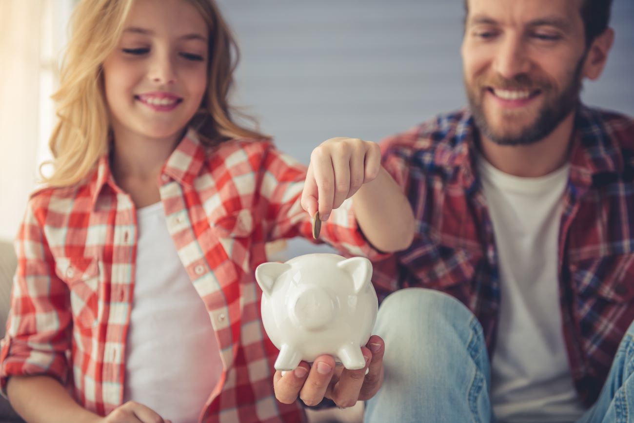 Smiling girl sits by smiling man both wearing checked shirts and puts a coin in a piggy bank