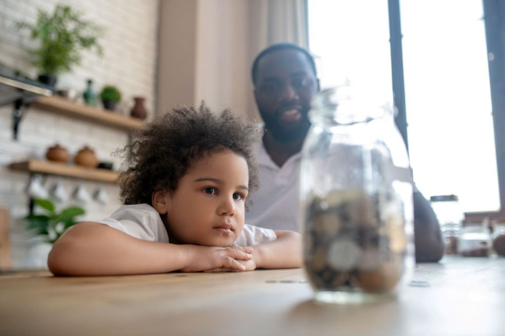 Young Black boy leaning on the table looking at jar of coins with Black man smiling in the background.