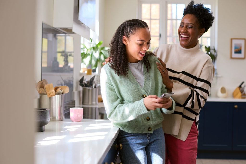 Teen girl holding phone stands next to woman who is smiling in kitchen. 