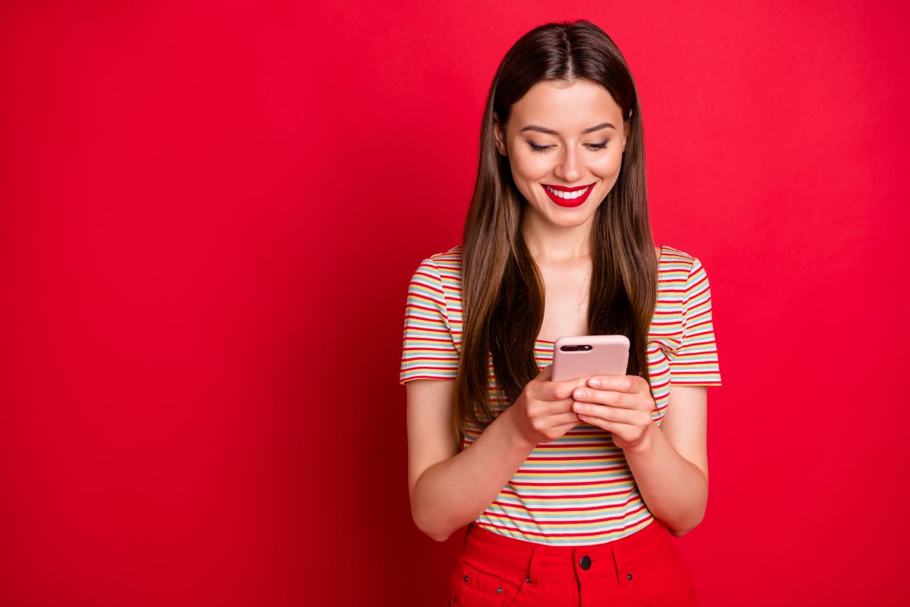 Girl wearing red stripped top holding smartphone and smiling stands against red background