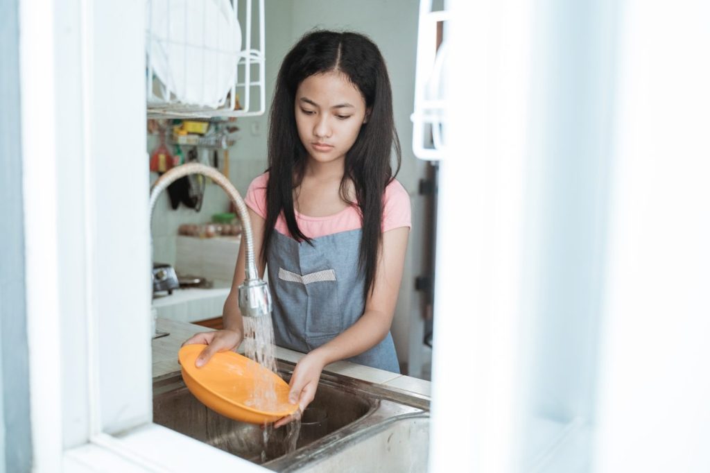 Teen girl standing at sink washing orange dish.