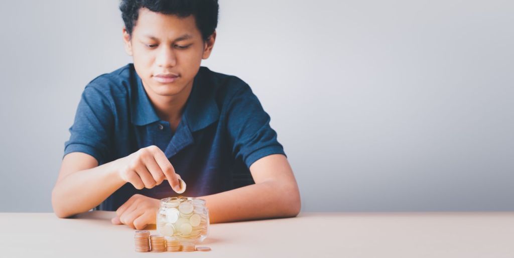 Boy in blue shirt puts coin in jar next to stacks of coins