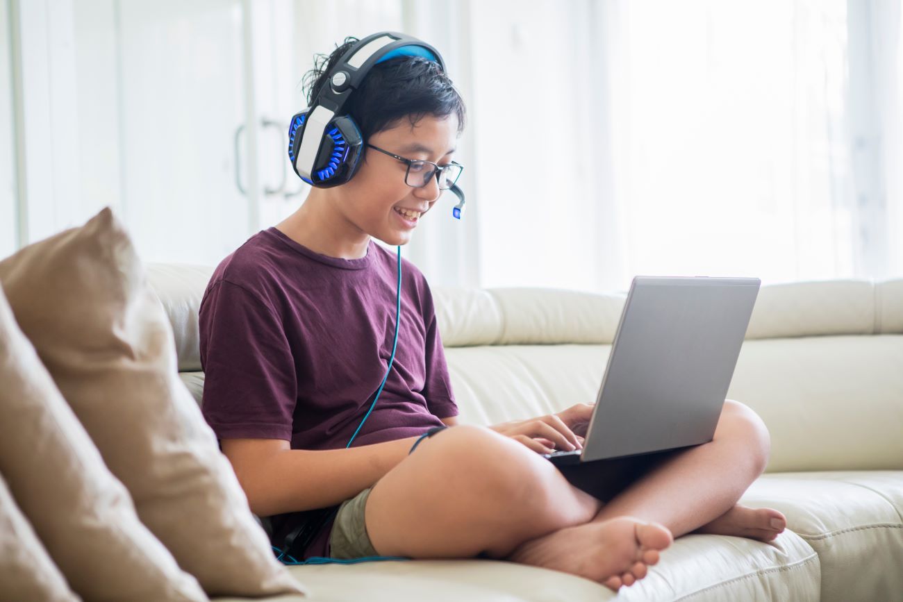 Boy sits on couch wearing headset playing game on laptop