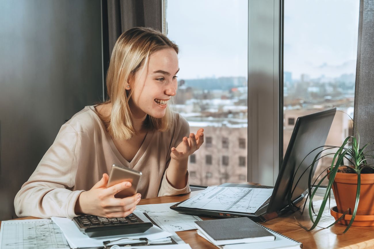 Young woman holding calculator looks at laptop surrounded by stock reports