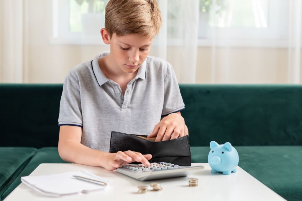 Boy inputs numbers into calculator and has pile of coins and piggy bank