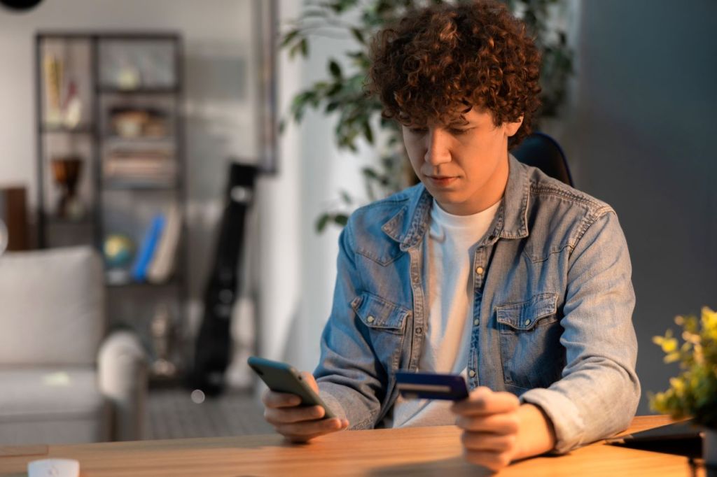 A teen boy holding a credit card and reviewing his bank statements on his phone