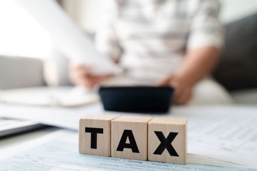 wooden blocks spell out "tax" with person in background using calculator