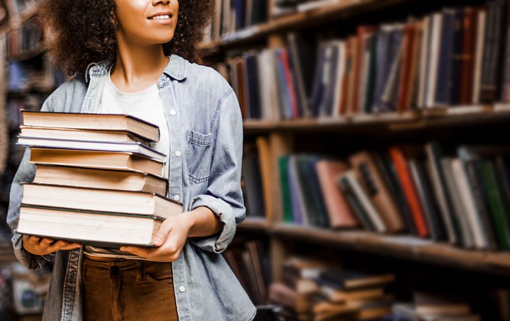 Teen girl holding pile of books and working at library