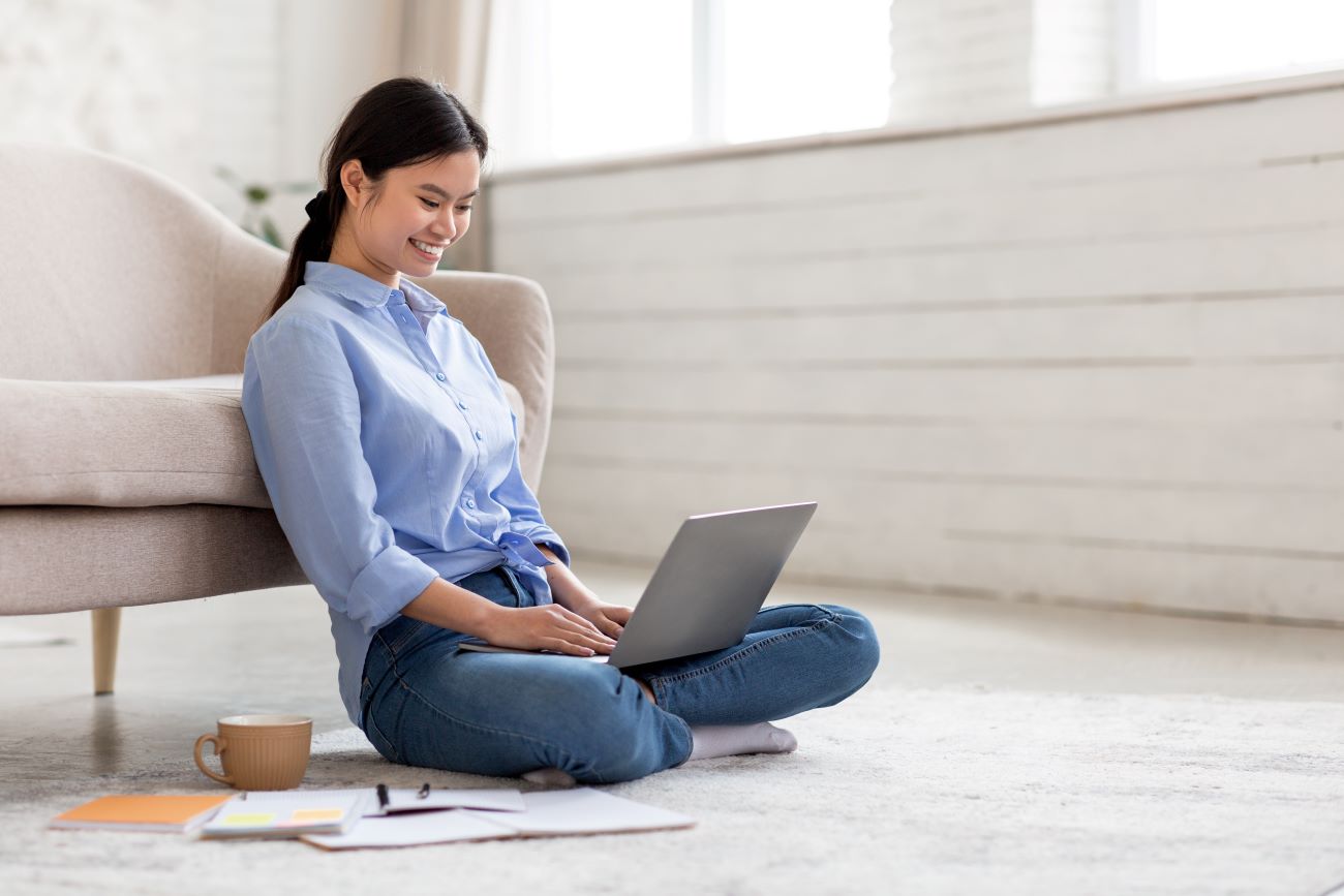 Smiling Asian teen girl sits on floor with laptop writing cover letter