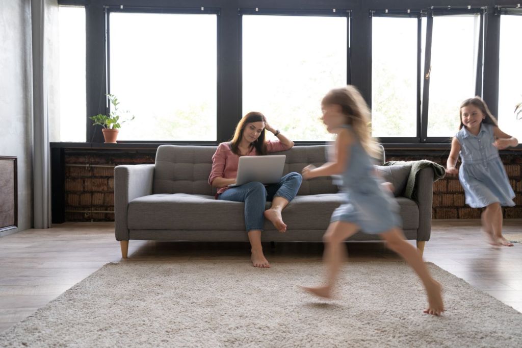 female coparent sits on couch with laptop calculating finances kids run around
