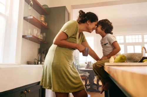 Single parent holding cup rubs noses with boy sitting on kitchen counter