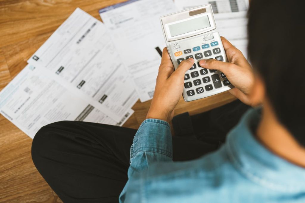 man holds calculator sitting on floor surrounded by bank statements