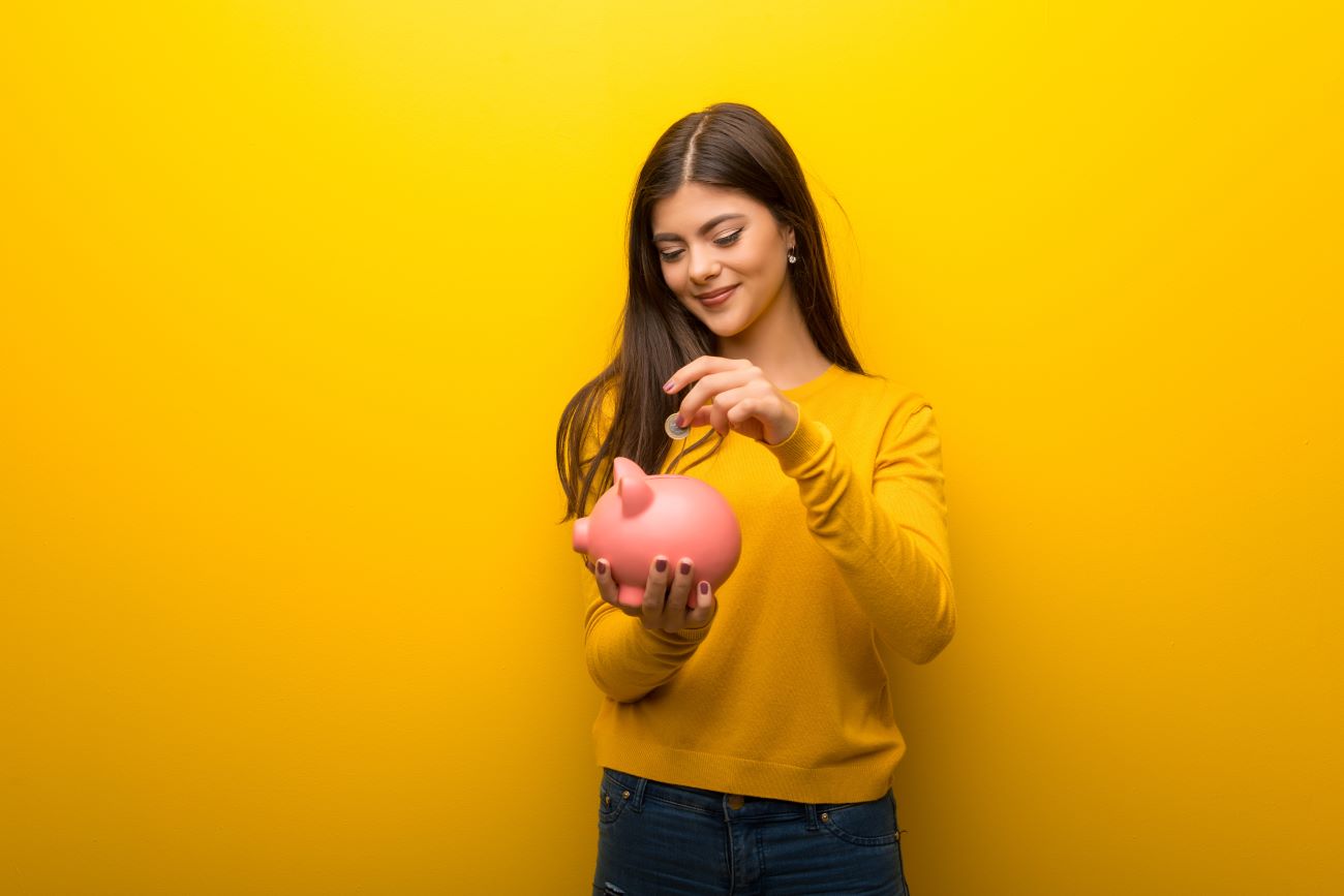 Girl wearing yellow sweater against yellow background puts coin in piggybank