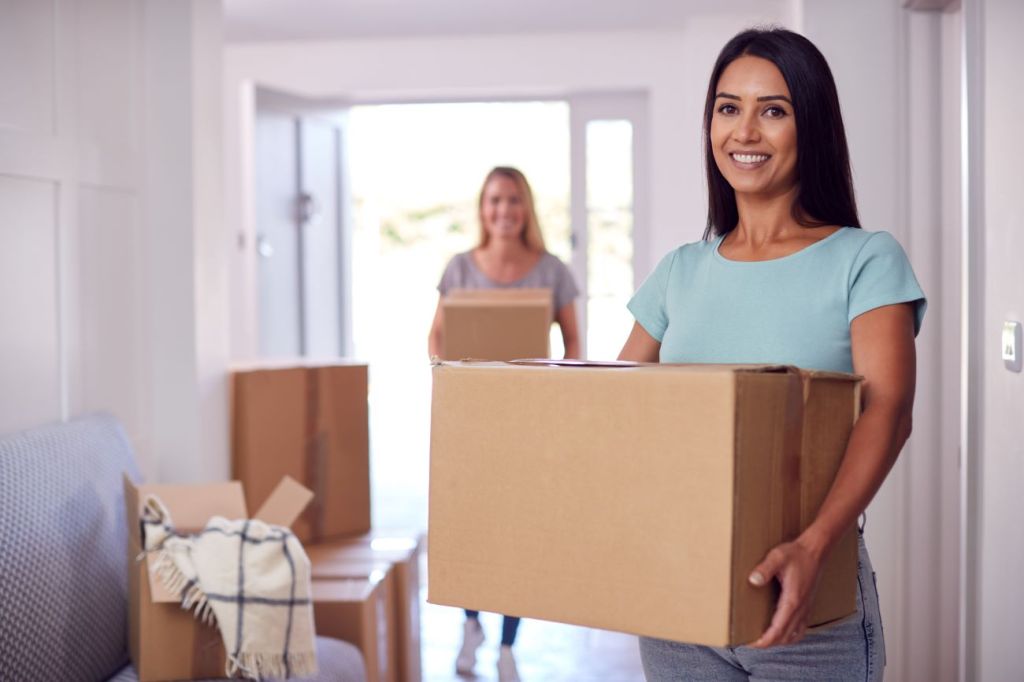 Two women smiling carrying moving boxes in their new home