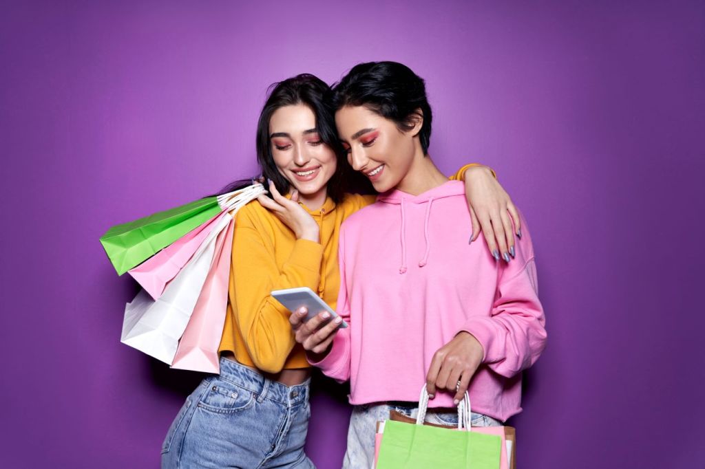 two young woman looking at phone holding shopping bags