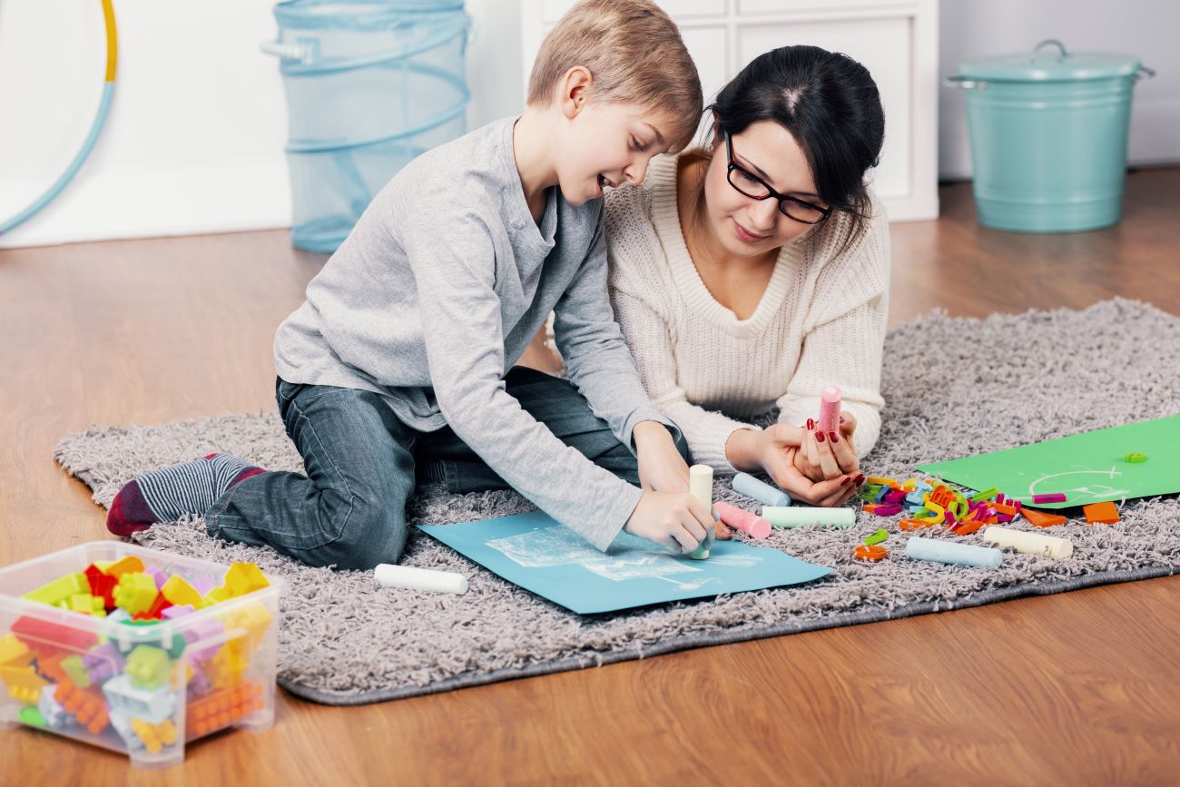 Boy with autism and his mom sit on floor he is drawing with chalk and she is looking on.