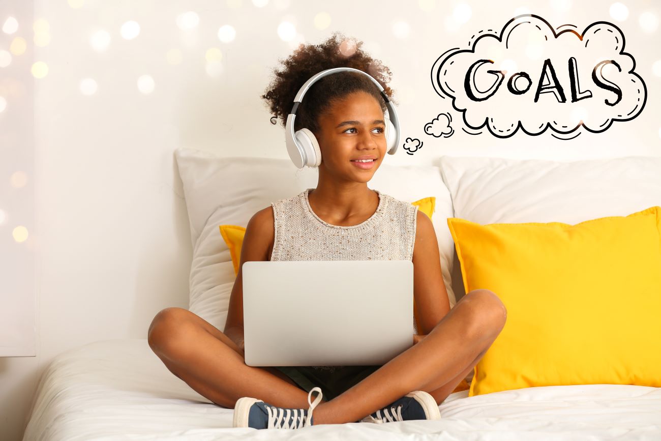 girl wearing headphones sitting on bed with laptop and word "goals" as thought bubble