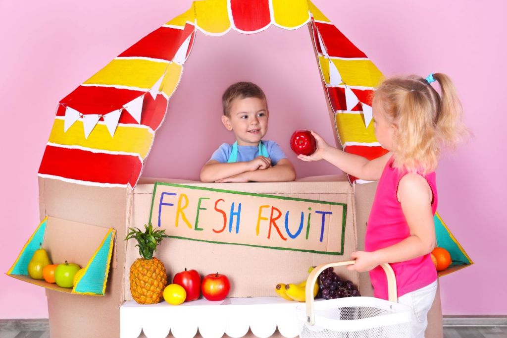 Young boy and girl standing at toy fresh fruit stand playing shop