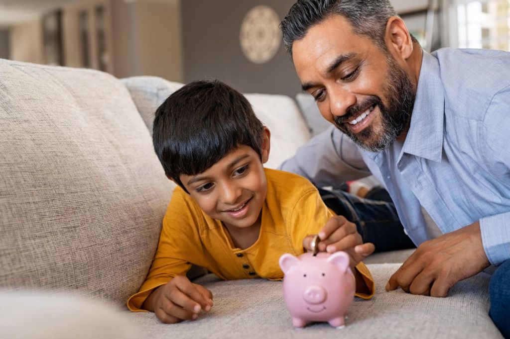 boy lies on couch putting coins in piggy bank with man looking on