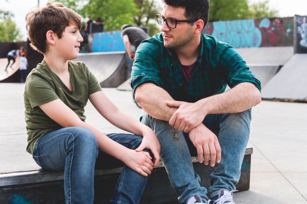 boy and man sit on ground at skating park talking about money