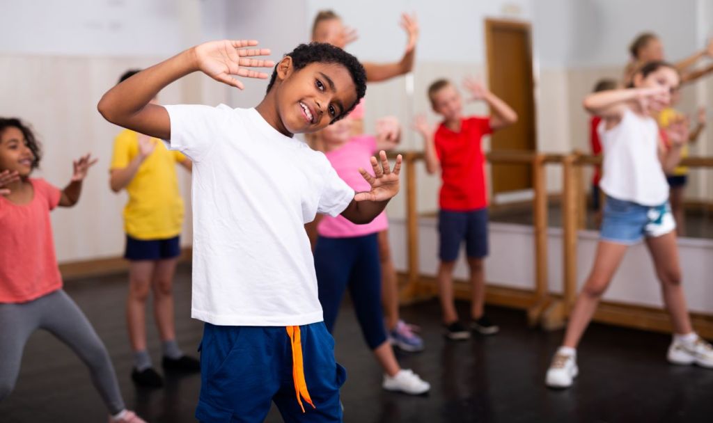 Boy wearing white t-shirt smiling with his hands in the air at a dance class with kids in the background