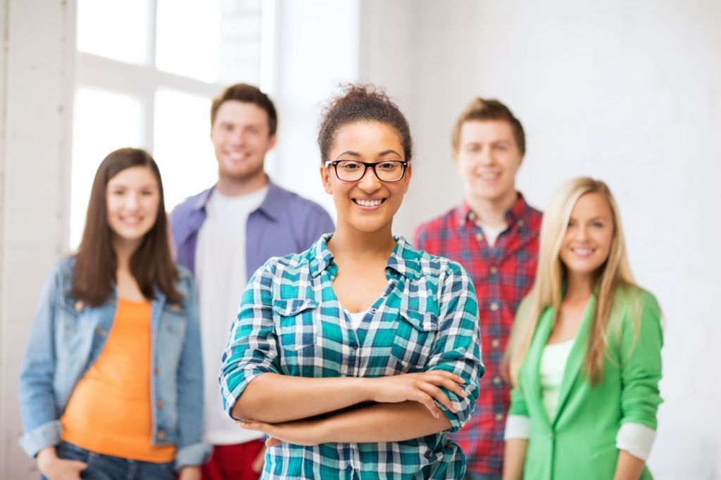 Smiling teen girl wearing glasses and arms folded stands in front of group of 4 teens