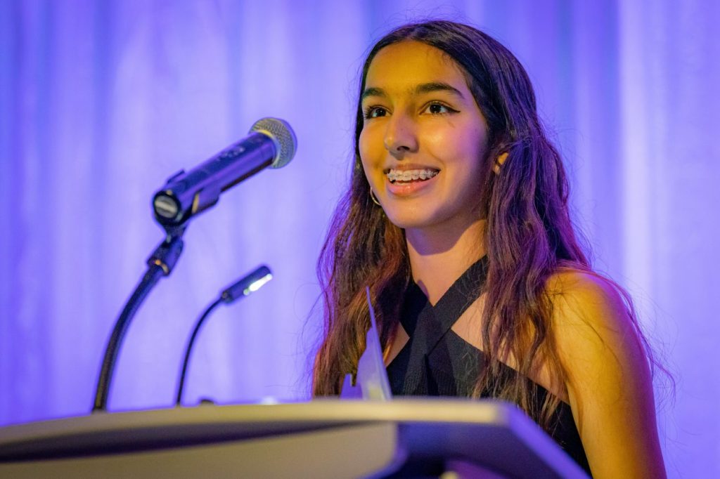 Image of Naila Moloo standing at podium speaking int a microphone with purple background.