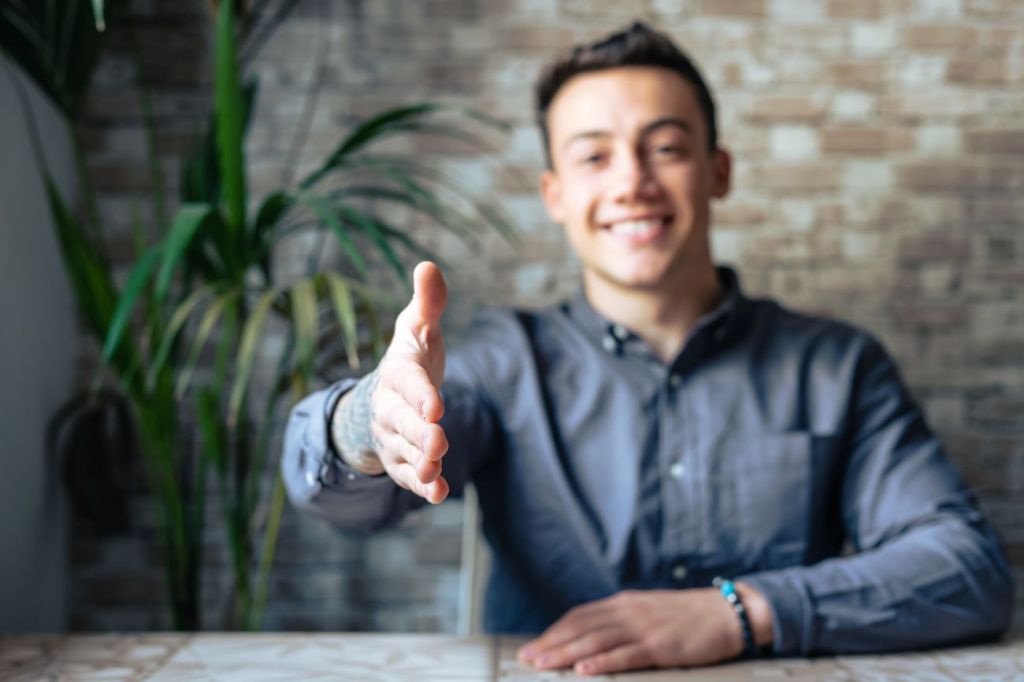 Smiling teen boy wearing shirt sits at desk and holds out hand to shake another.