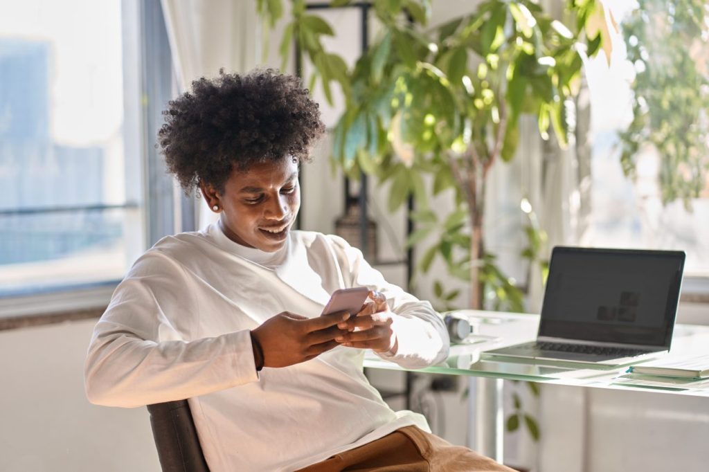 Black teen boy sitting at desk with laptop holding smartphone and smiling. 