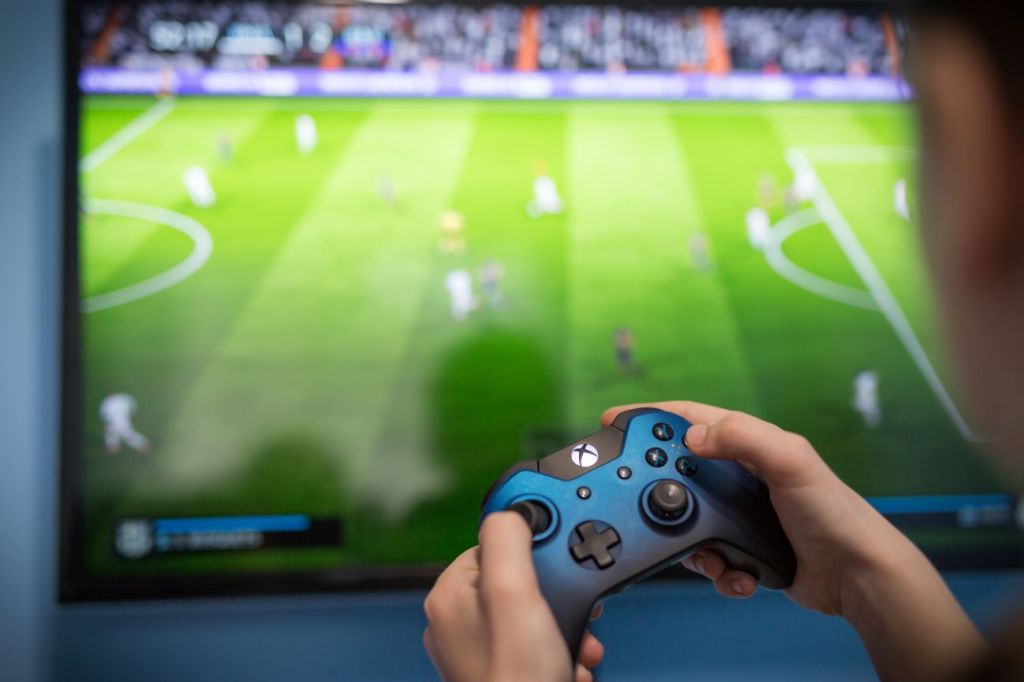 Image of hand holding blue video game controller playing FIFA on a screen in background.