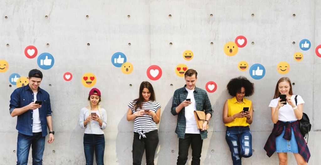 Group of teens on their phones standing against a grey wall with social media emojis above their head