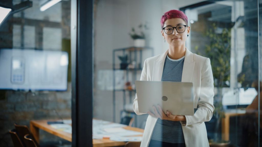 LGBTQ woman with short pink hair wearing white jacket holding laptop in the workplace