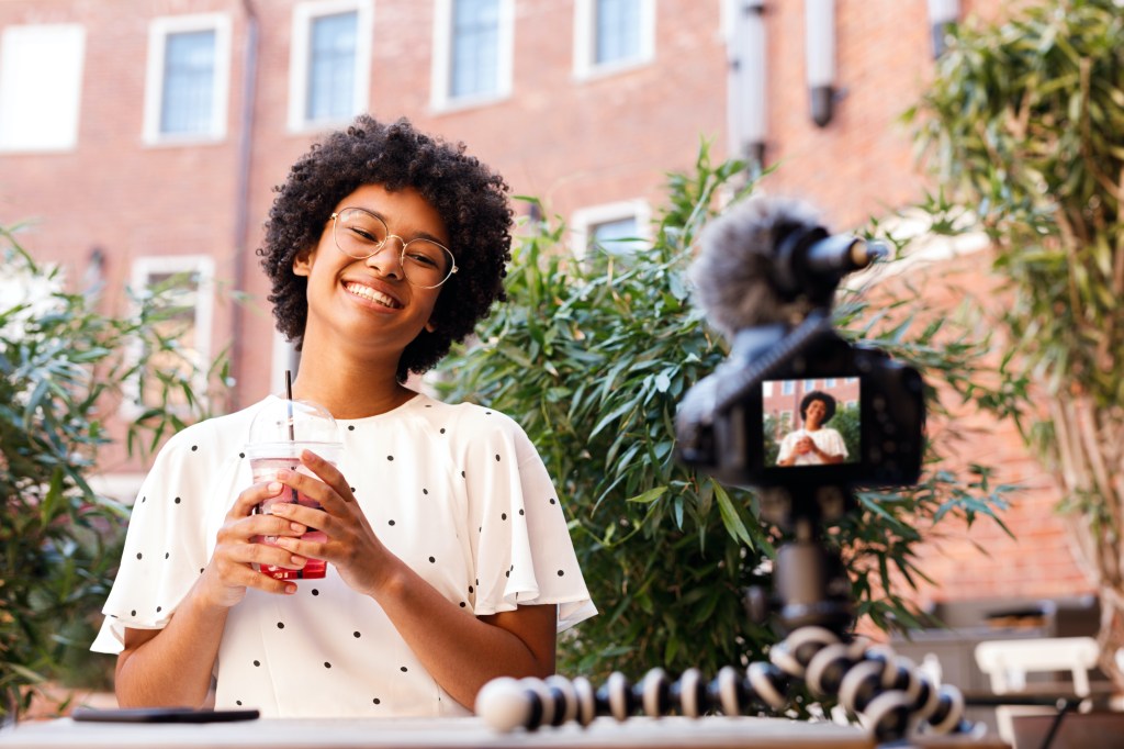 Happy teen girl who is a content creator recording a video on dslr camera, holding a juice