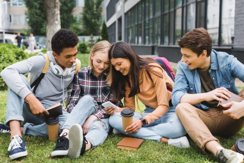 Group of smiling teens sitting on grass looking at phone and drinking coffee