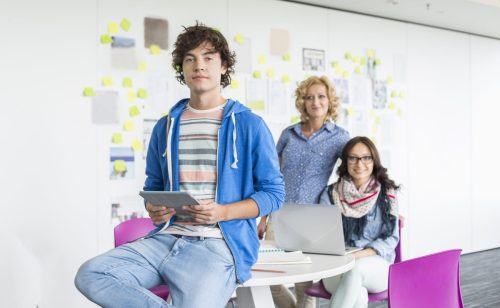 Male teen intern sitting and holding ipad with two women colleagues in the background