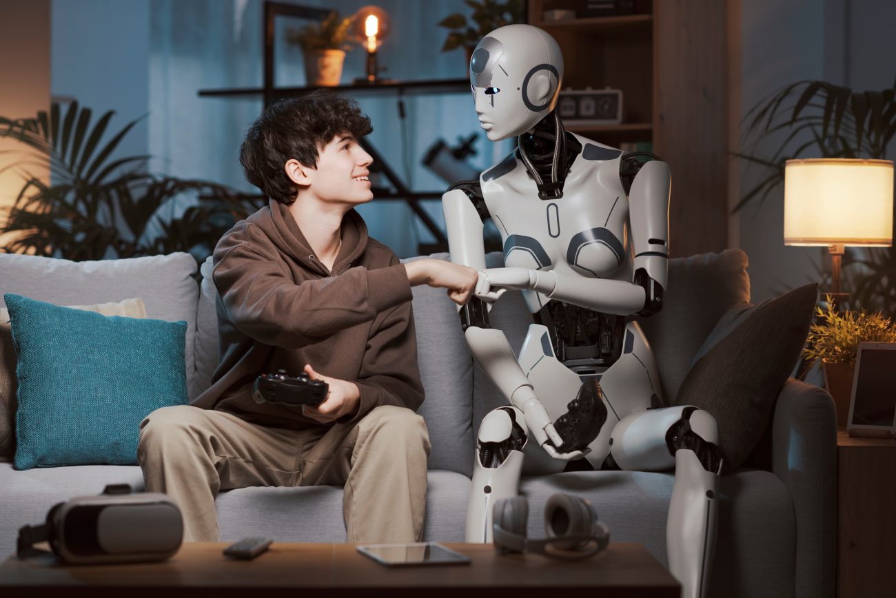 Smiling teen boy sits on couch looking at robot and fist bumping it.