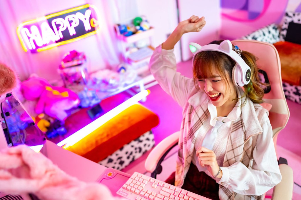 Asian teen girl wearing pink headphones sits at computer in colourful bedroom playing video games