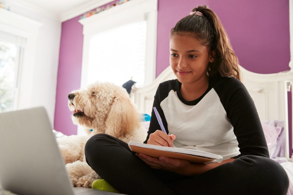 Smiling teen girl sitting on bed doing homework next to a white dog