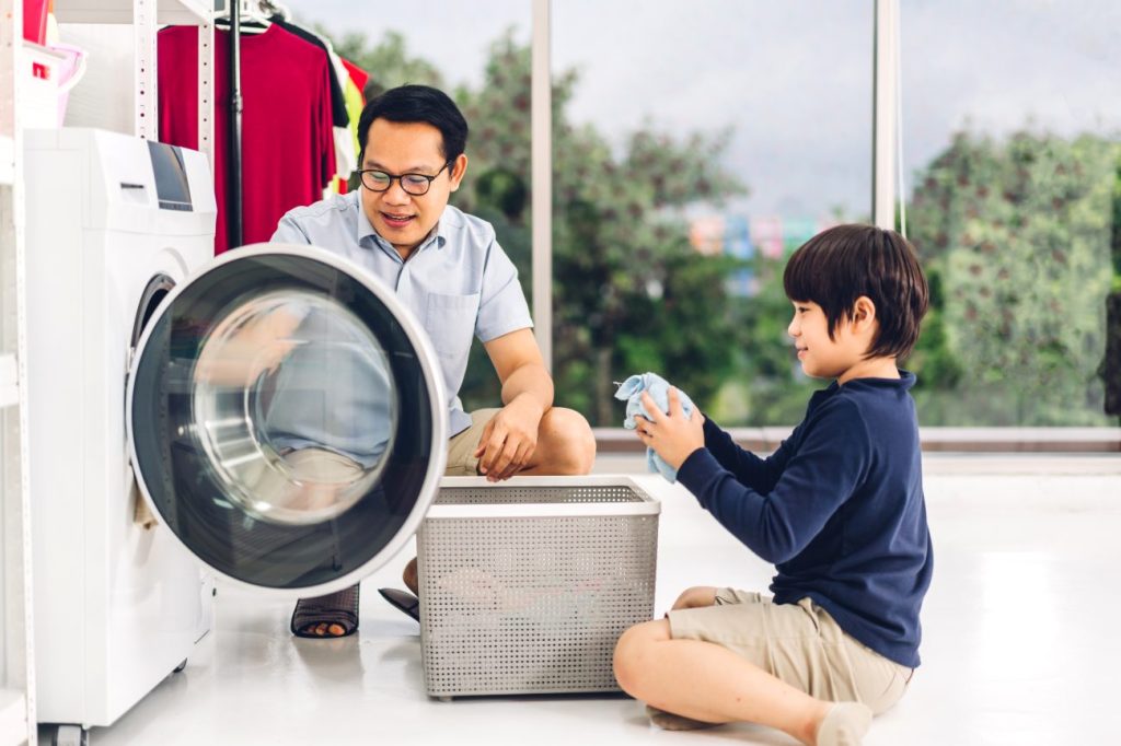 Man and boy sitting on floor sorting out laundry and putting it in washing machine. 