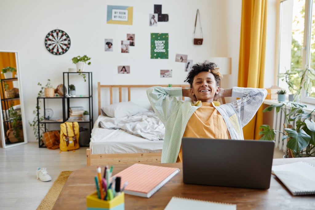 Black teen boy leaning back in decorated bedroom at his desk with wall art in the background
