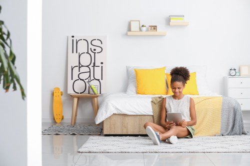 Black teen girl sits on floor next to bed with yellow pillows and artwork saying "inspiration" in background