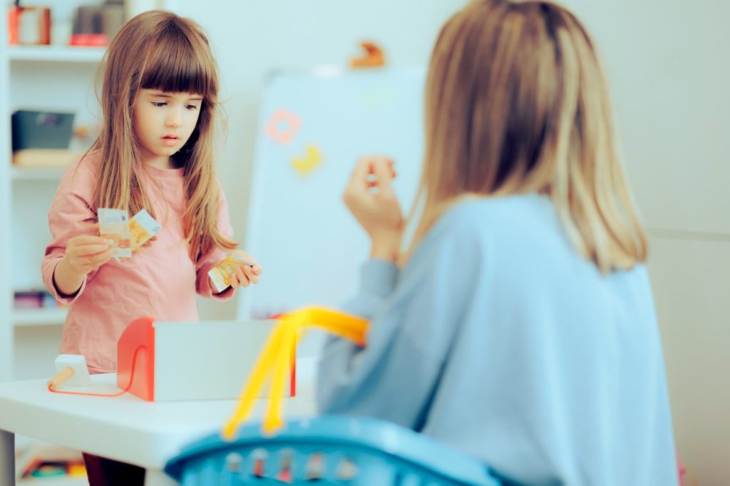 Little girl playing with woman holds play money and stands over toy cash register.