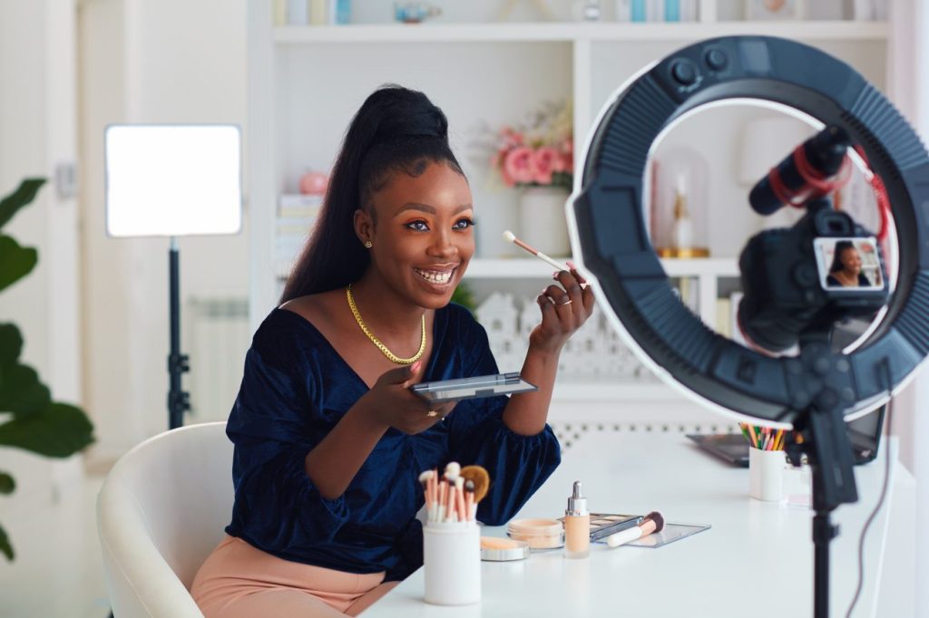Smiling Black woman demonstrates how to put on makeup in front of ring light