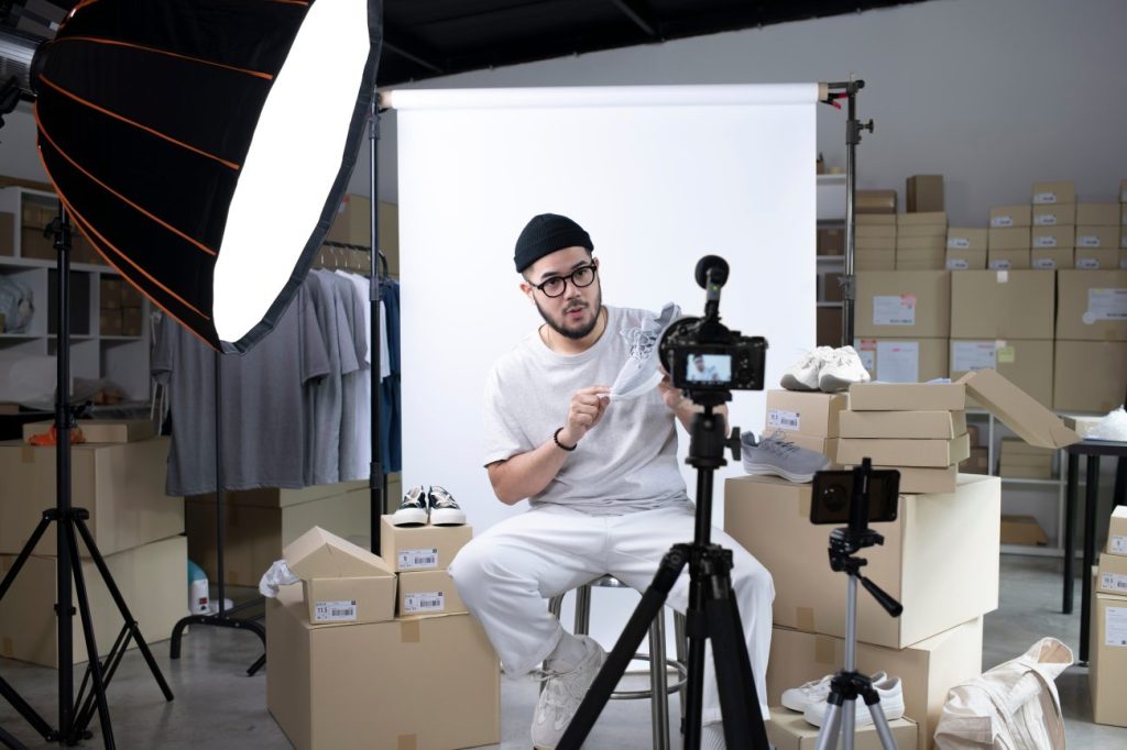 Asian man sitting on chair with white screen behind him showing sneakers to camera.