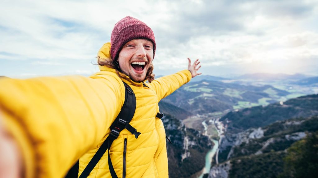 Smiling male travel influencer wearing yellow coat stands on top of mountain