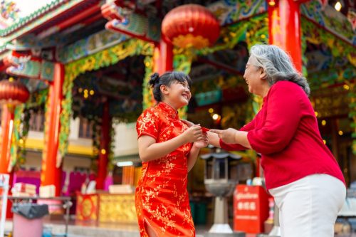 Smiling Asian Girl wearing red dress accepts lucky red envelope from smiling older Asian lady