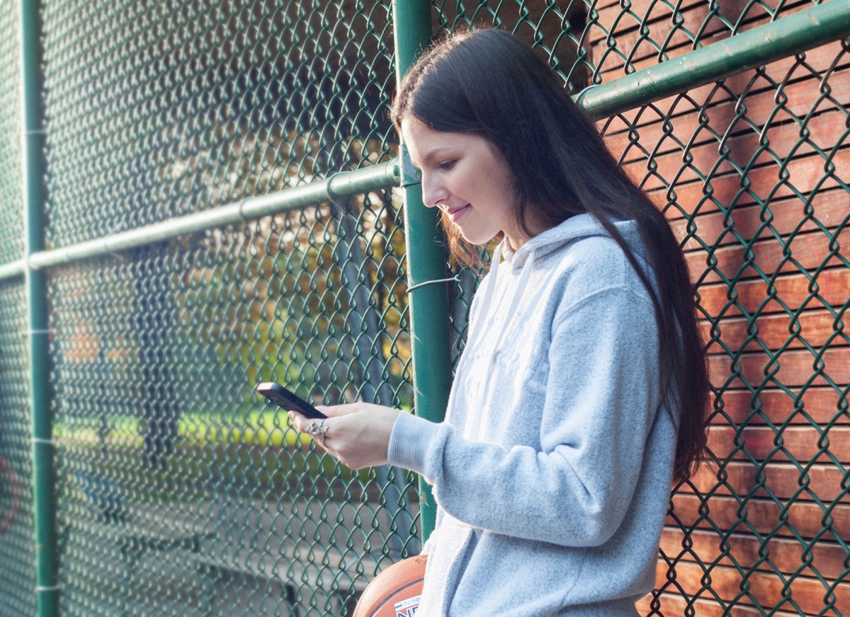 A young person in a gray hoodie leans on a fence holding a basketball and a mobile phone, showing how it works to manage money.