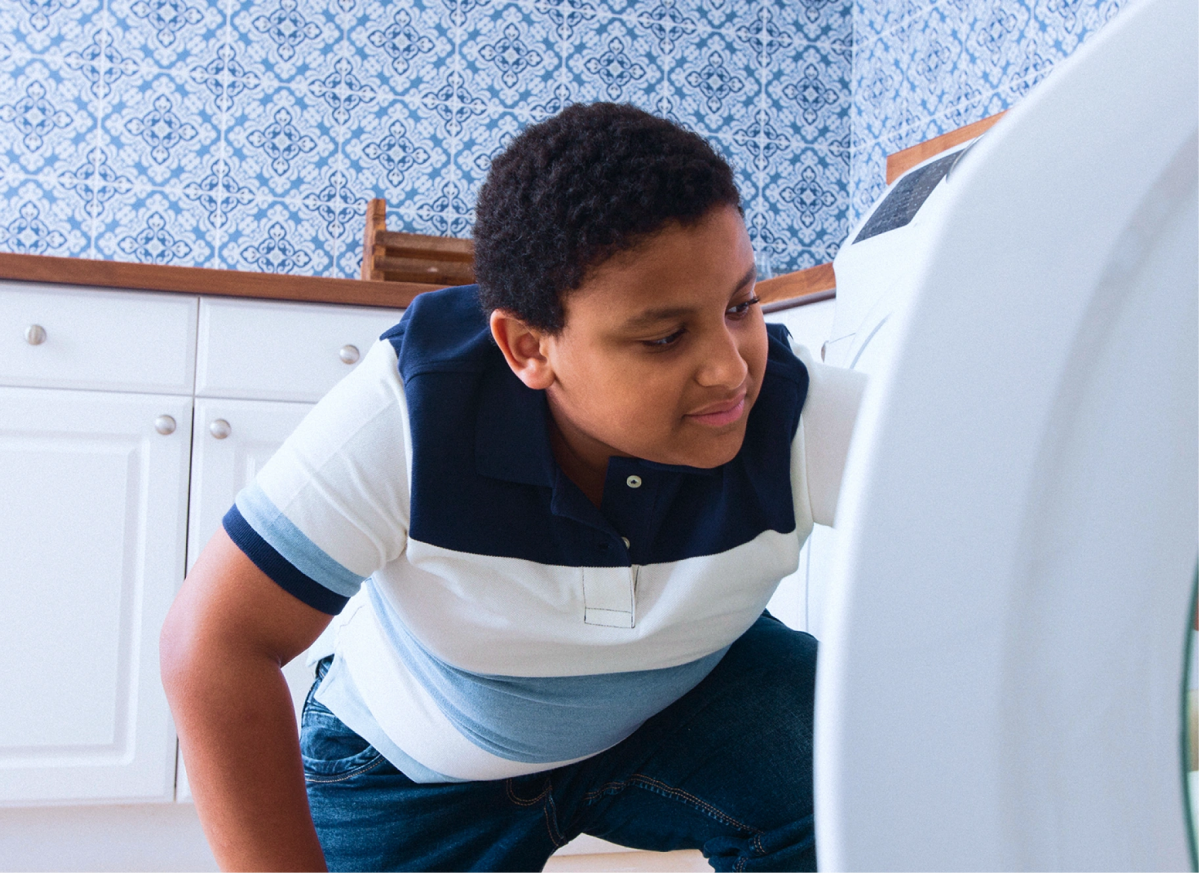 A young person crouches by a front-loading washing machine, showing how it works to handle chores and money at home.
