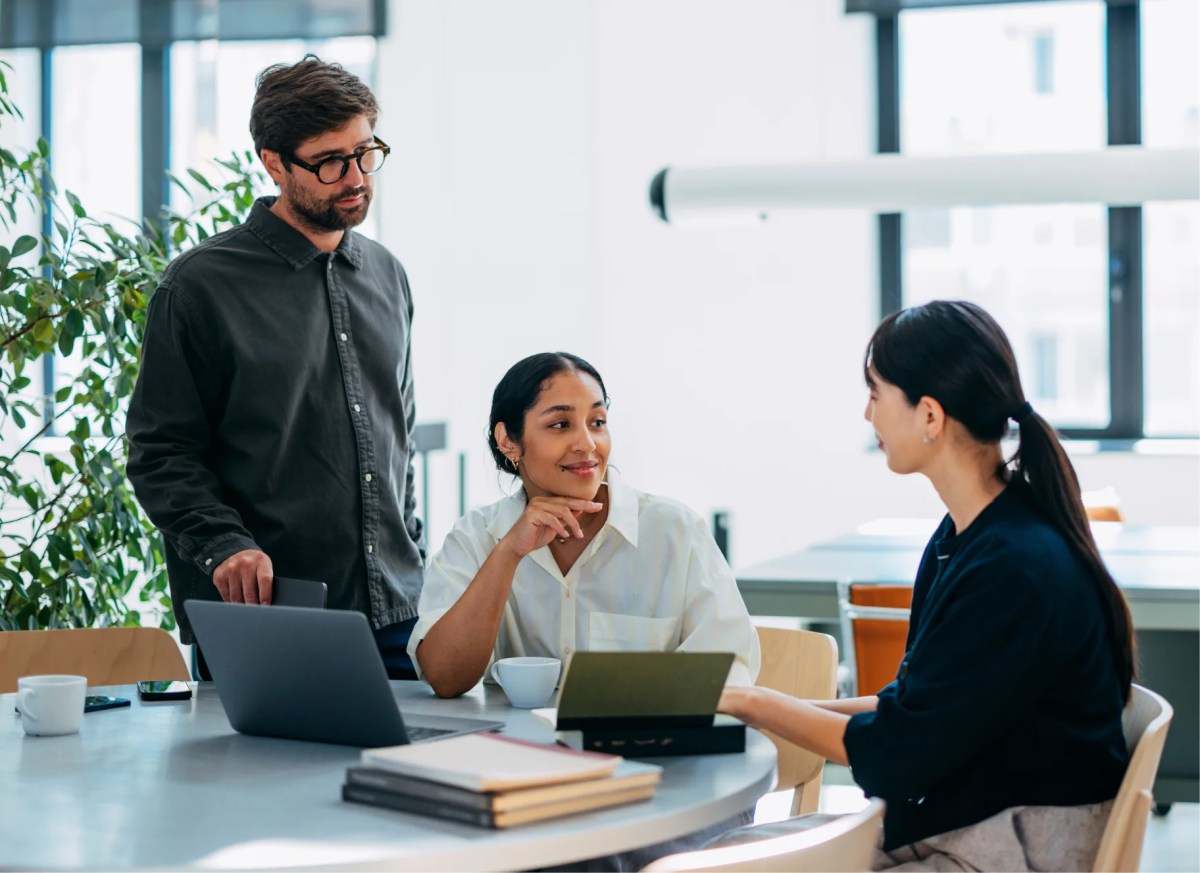 Three colleagues collaborating around a round table in a bright modern office, discussing ideas over laptops and notebooks during a career meeting.